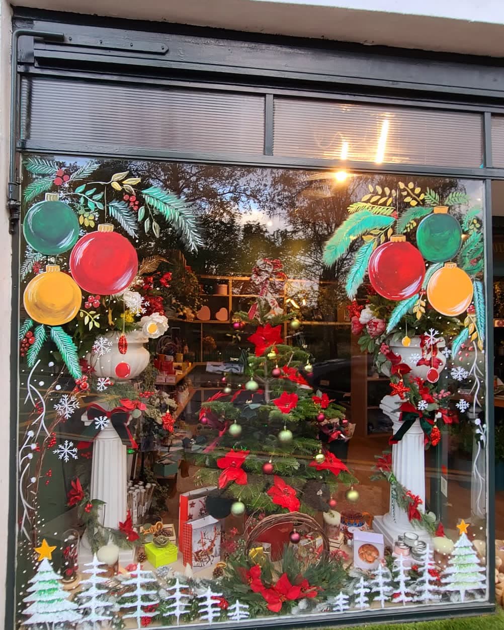 A florist's window hand-painted for Christmas — green, red and yellow baubles hung from teal pine sprays and gold leaf sprigs, white snowflakes scattered down the glass and a row of white fir trees along the bottom, with the shop's own poinsettias and Christmas tree showing through behind.